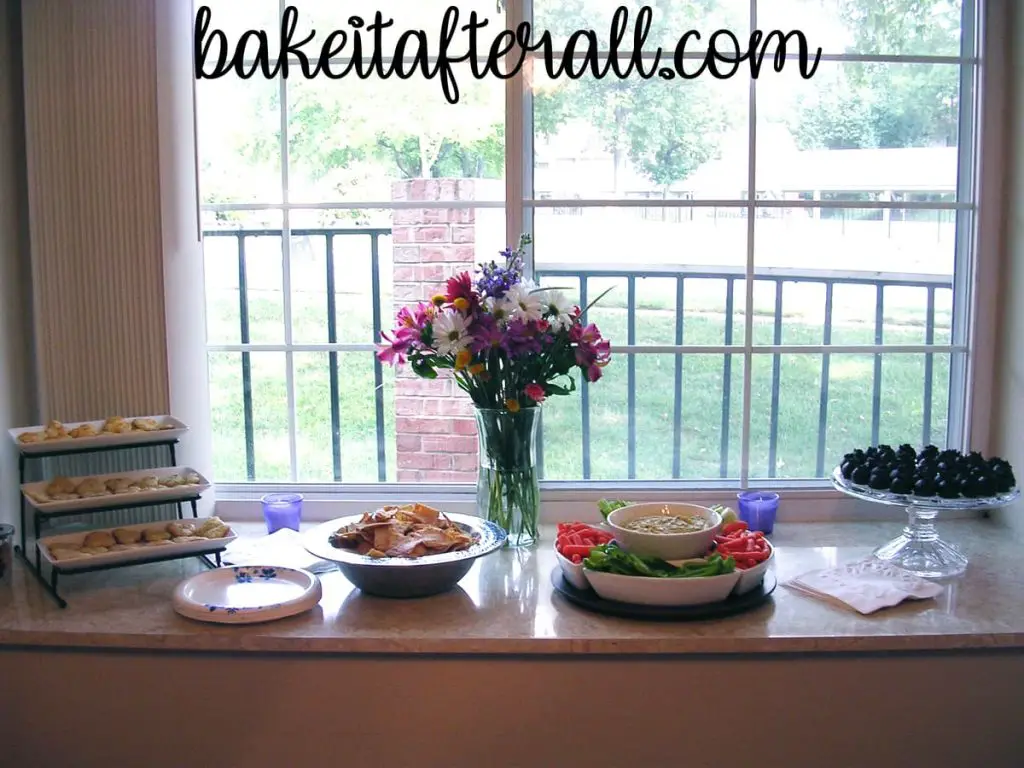 food and desserts on a counter with a vase of flowers in the center in front of a window