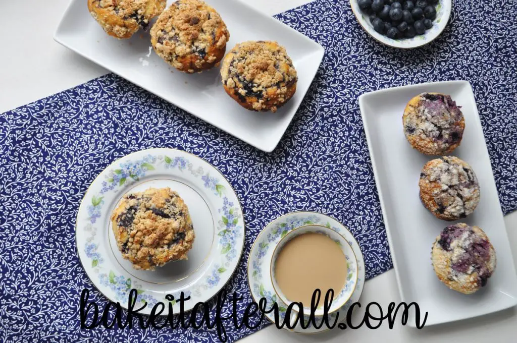 overhead shot of best blueberry muffins on white platters with a blue and white flowered table runner and a cup of coffee