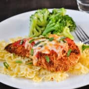 Baked Chicken Parmesan on top of pasta on a white plate with broccoli in the background