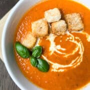 a bowl of creamy tomato soup with a swirl of cream, croutons and fresh basil leaves on top