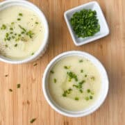 two white ramekins of leek and potato soup on a wooden cutting board sprinkled with chopped chives
