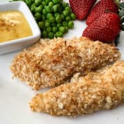 Coconut Crusted Chicken Tenders on a white plate with peas, strawberries and a small dish of honey mustard