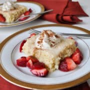 a piece of tres leches cake on a gold-rimmed China plate surrounded by fresh strawberries with whipped cream and cinnamon on top