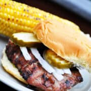 a turkey burger on a bun with pickles and onions with an ear of corn on the cob behind it