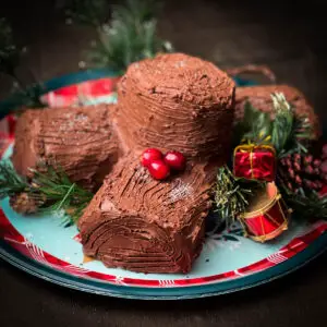 a Christmas yule log buche de noel on a platter decorated with sprigs of evergreen and holly berries