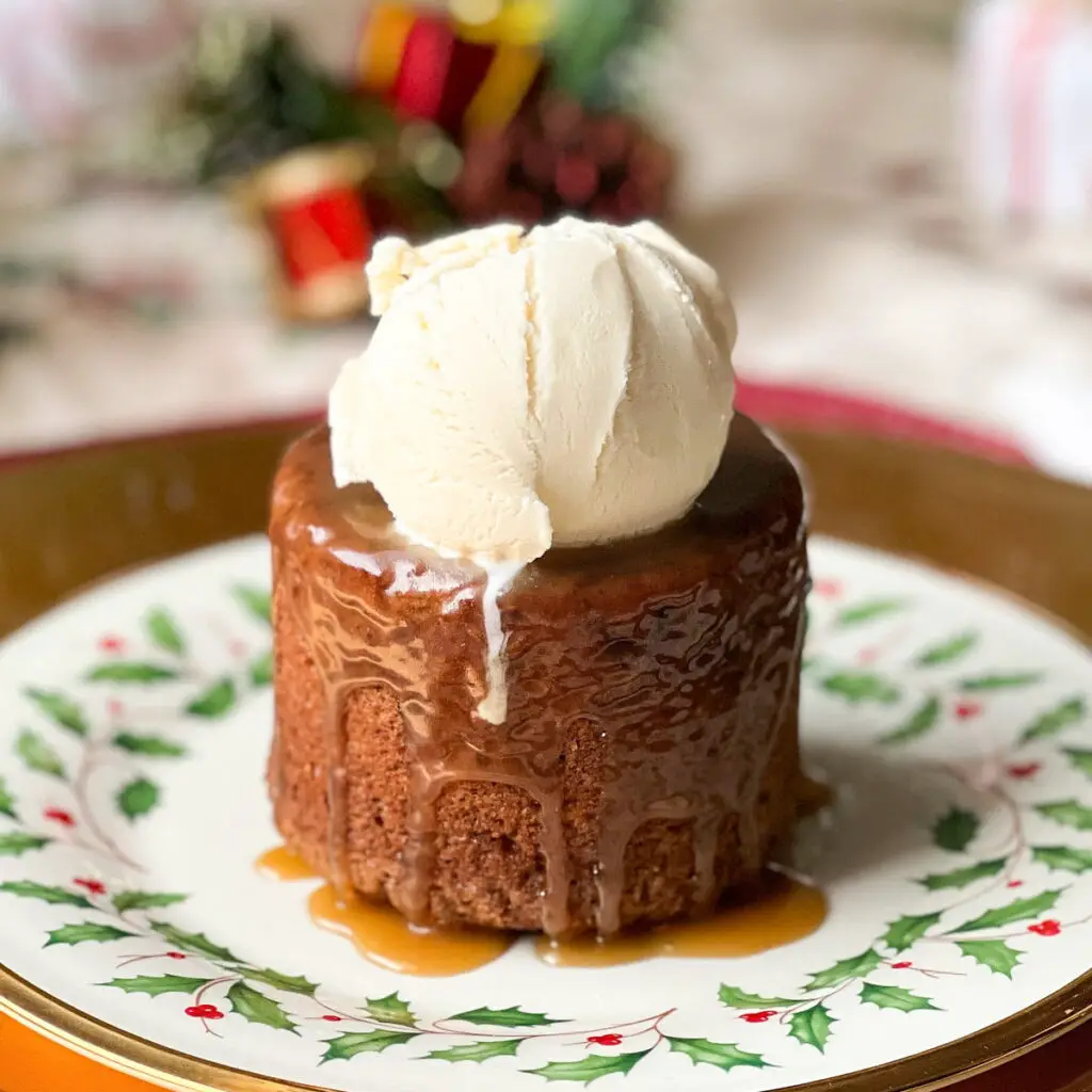 Warm sticky figgy pudding on a holly plate topped with caramel sauce and a large scoop of vanilla ice cream