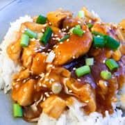 rice on a plate covered with skinny orange chicken that has been sprinkled with fresh green onions and sesame seeds
