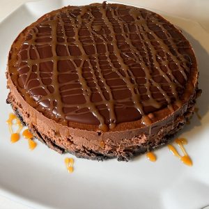 Gluten Free Mocha Cheesecake topped with dark chocolate ganache drizzled with caramel sauce on a white cake stand