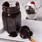 ben & jerry's homemade hot fudge in a ladle on a white plate next to a jar of sauce with a hot fudge sundae in the background