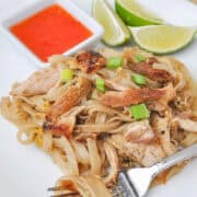 chicken pad thai on a white plate with a fork and wedges of limes