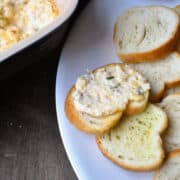 Asiago cheese dip in a casserole dish next to a plate of crostini with dip spread on the top one