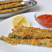 baked zucchini sticks on a white plate with a cup of marinara sauce and some lemon wedges