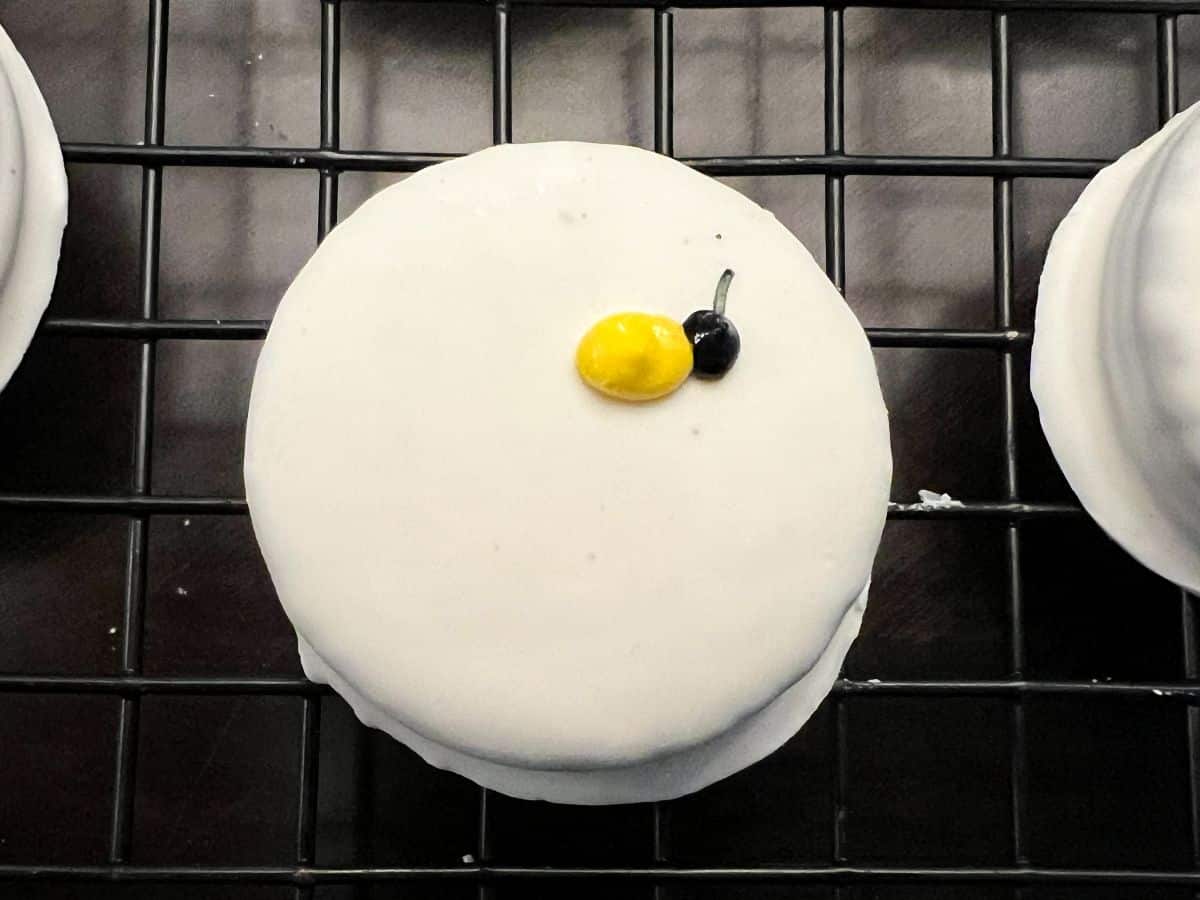 a yellow oval of royal icing next to a black dot of royal icing on a white fudge covered Oreo