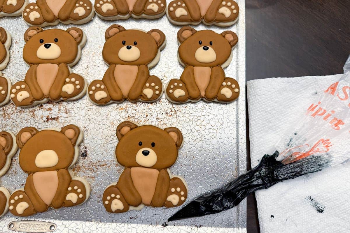 finished royal icing teddy bear cookies on a cookie sheet next to a piping bag filled with black royal icing