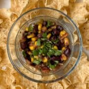 Adobo black bean and corn salsa in a glass bowl with tortilla chips around it