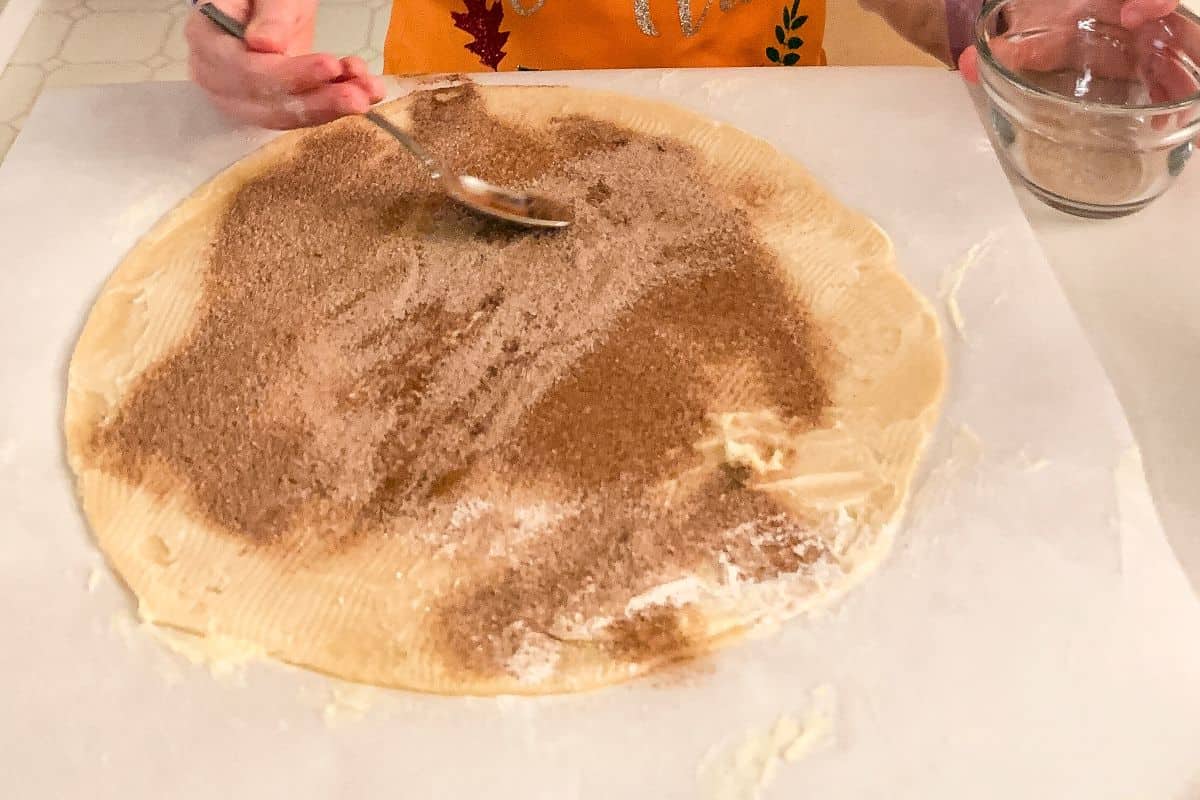 a child's hand spreading cinnamon sugar over a rolled pie dough using a spoon