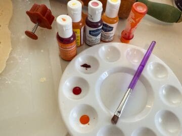 a paint palette with wells filled with food coloring gel, topped with a food grade paintbrush, next to bottles of food coloring gel
