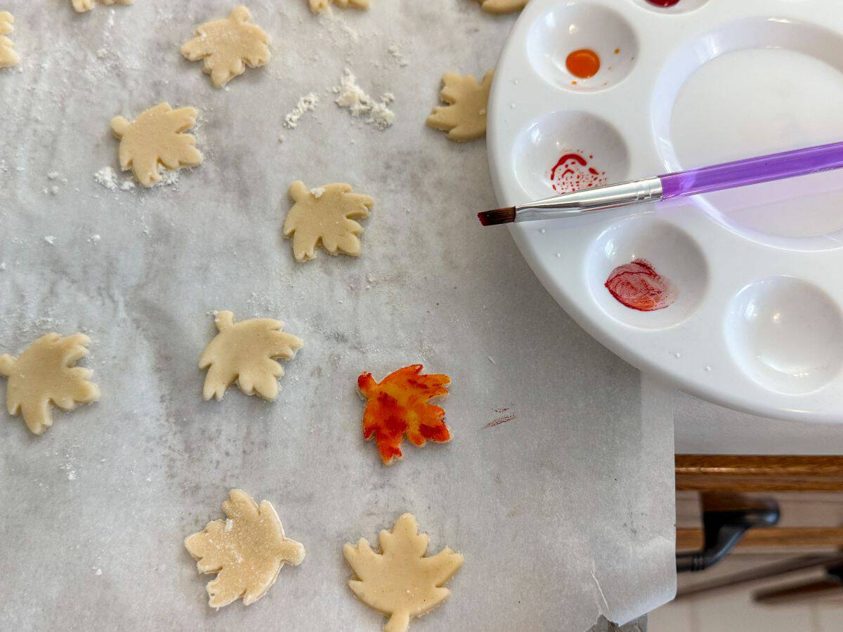 pie crust leaves on a parchment lined cookie sheet next to a paint palette with food coloring gels in the wells and a food grade paintbrush