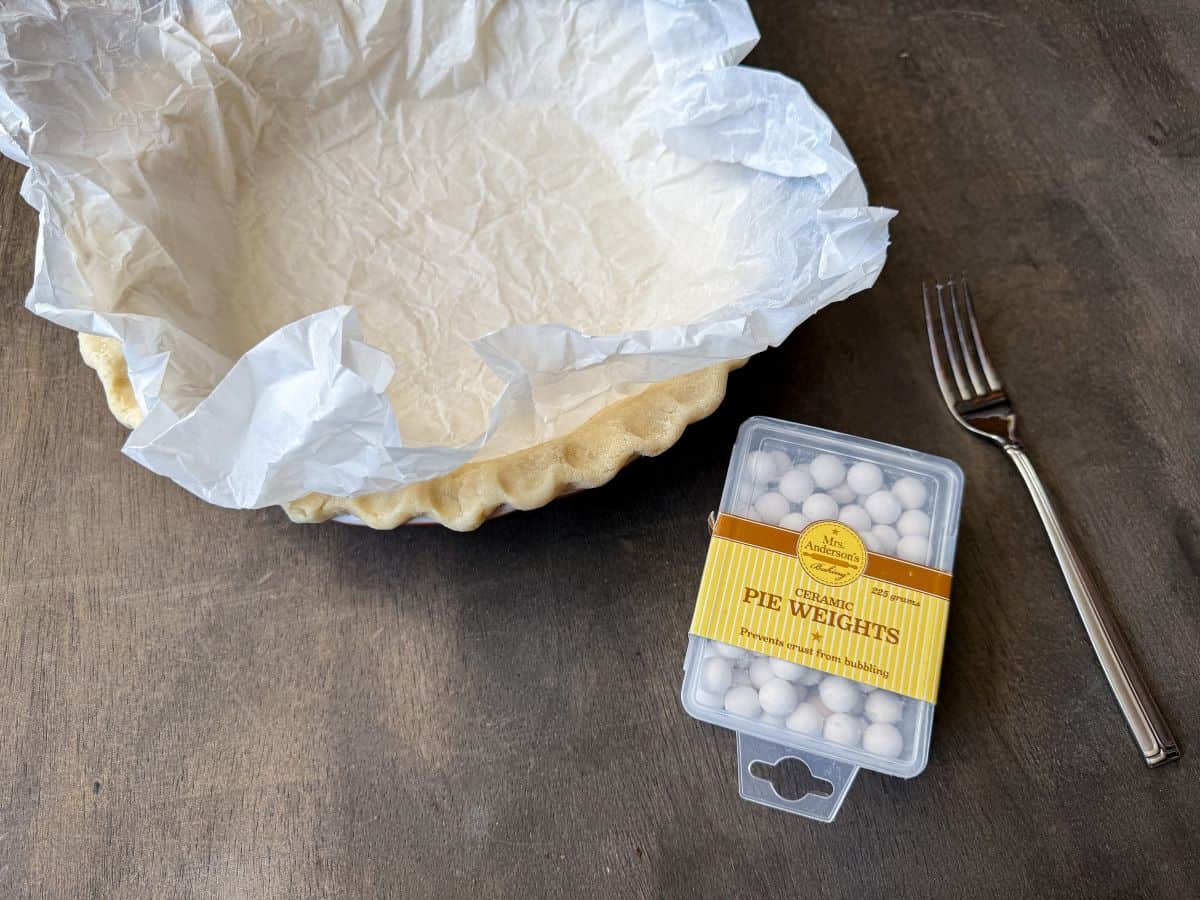 pie dough in a pie plate with crumbled parchment paper lining it next to a container of pie weights