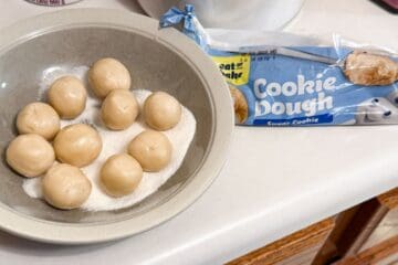 balls of refrigerated sugar cookie dough in a bowl of granulated sugar next to the package
