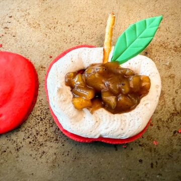 the bottom shell of a red apple macaron with an apple cider buttercream dam filled with caramel apple filling and a small piece of pretzel stick to represent a stem with a green fondant leaf