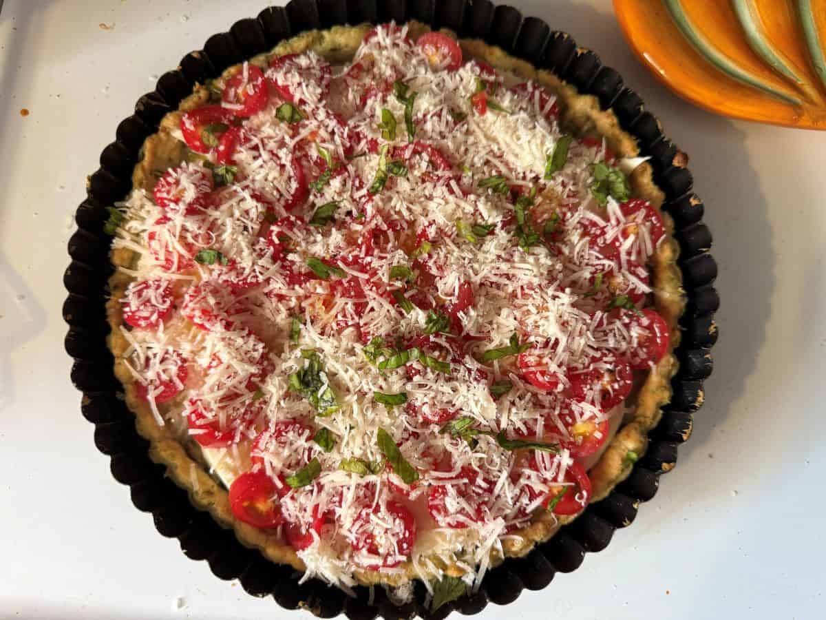 a tart pan filled with crust, cheese, sliced cherry tomatoes and covered in parmesan cheese and fresh basil
