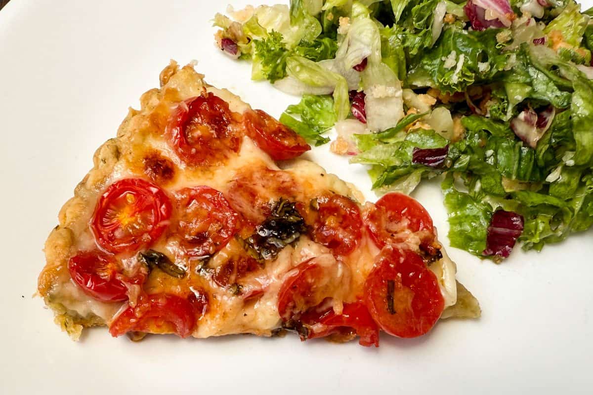 a slice of tomato mozzarella tart on a white plate next to a side salad