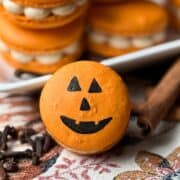 jack-o-lantern macaron on a fall kitchen towel with cinnamon sticks and cloves around it