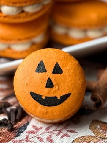 jack-o-lantern macaron on a fall kitchen towel with cinnamon sticks and cloves around it