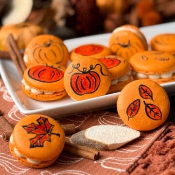 Painted Fall Macarons on a white plate with an orange fall kitchen towel, a wood slice, and fall foliage