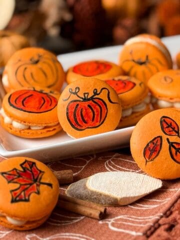 Painted Fall Macarons on a white plate with an orange fall kitchen towel, a wood slice, and fall foliage