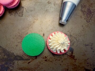 vanilla buttercream piped on top of a pink macaron shell next to a green shell