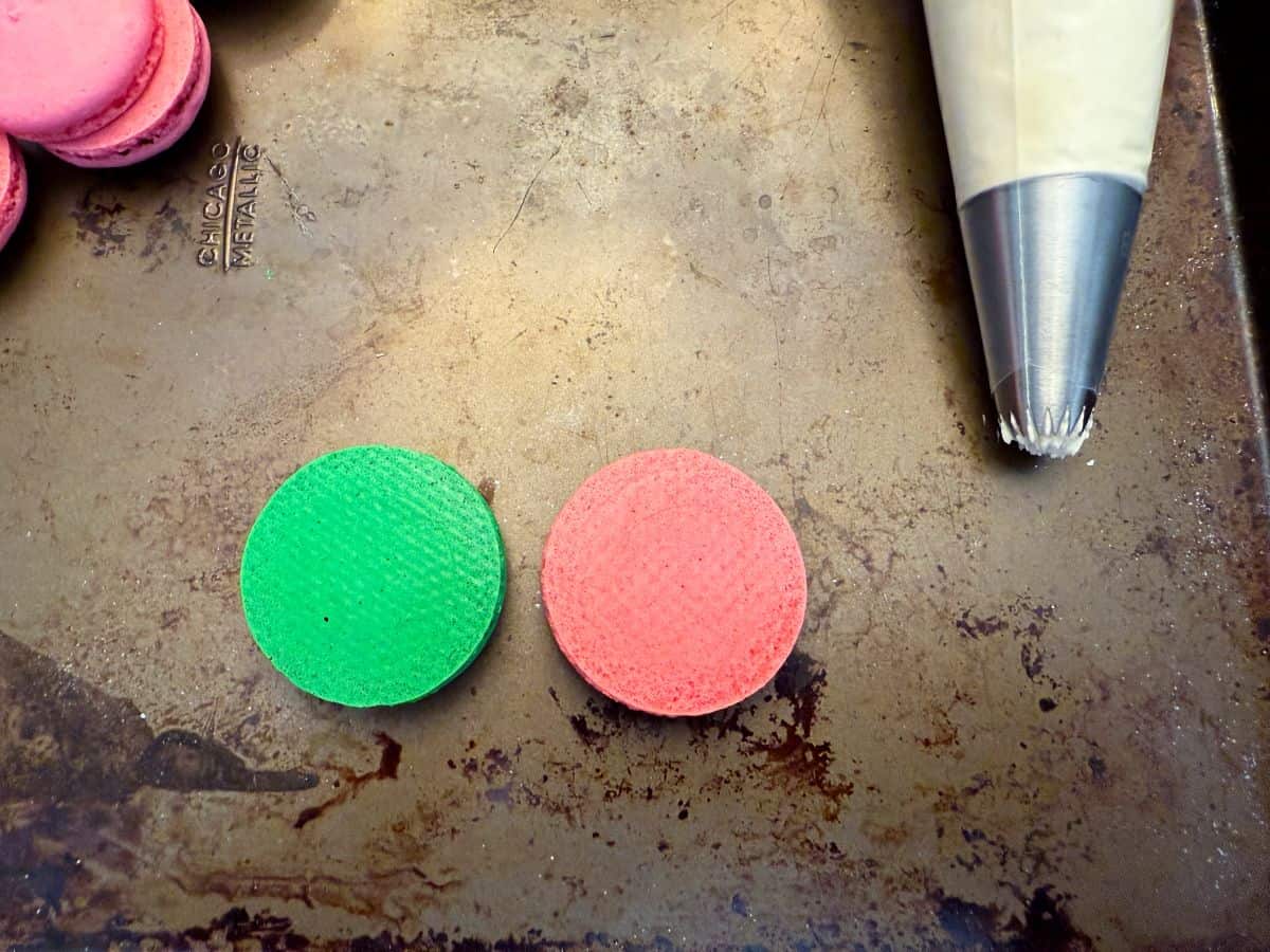 a green macaron shell next to a pink shell and a pastry bag of buttercream