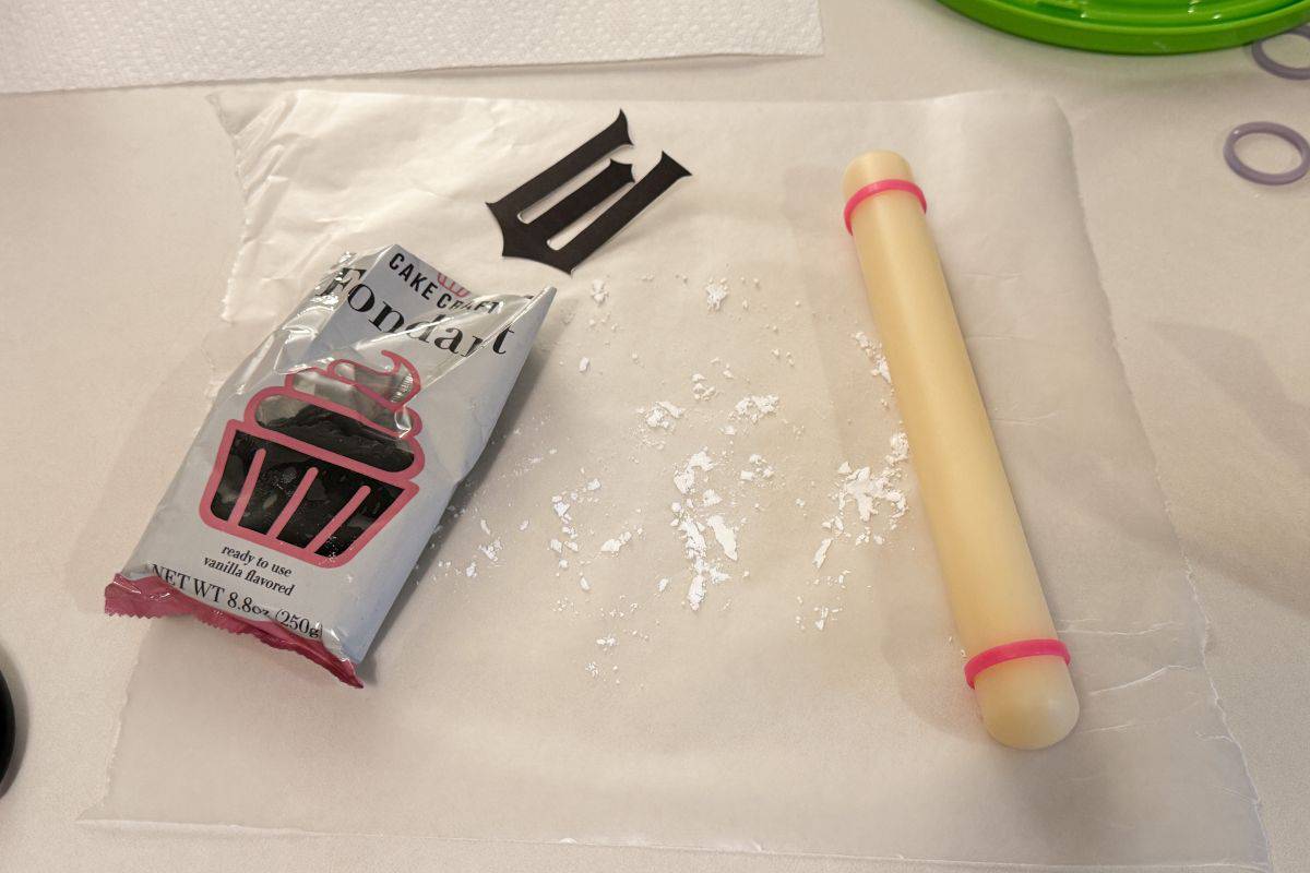 a piece of waxed paper with a package of black fondant, a black W cut out and a small fondant rolling pin