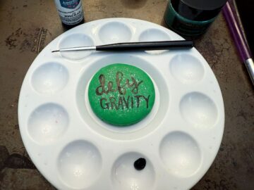 a green macaron in the middle of a paint palette with "defy gravity" painted on in black food coloring gel