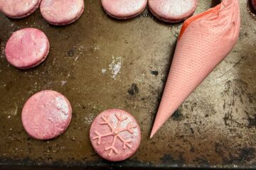 a pink macaron with a piping bag of raspberry buttercream next to it and a snowflake piped on top