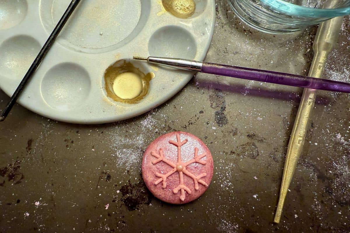 a paint palette with a well of edible gold luster dust suspension next to a macaron with a snowflake piped on top