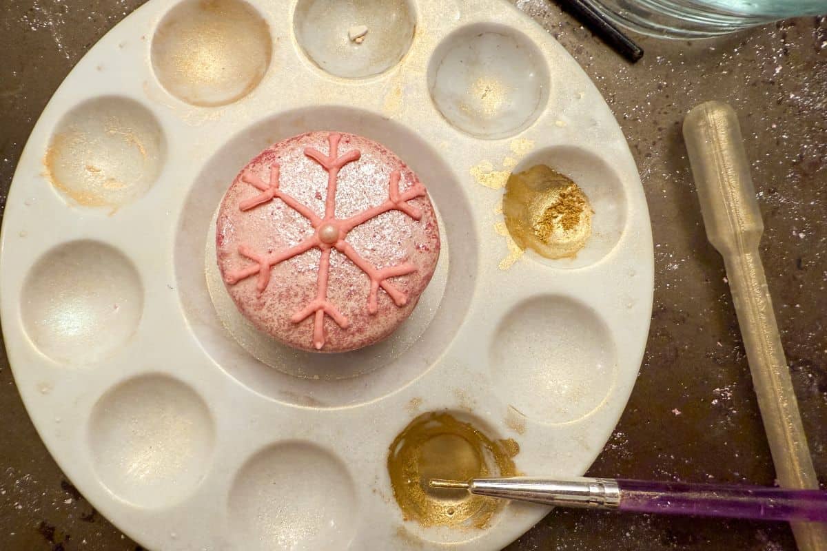 a pink macaron with a pink buttercream snowflake piped on top in the center of a paint palette and wells filled with gold luster dust and a paintbrush in the liquid