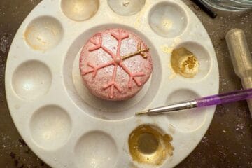 a pink macaron with a pink buttercream snowflake piped on top in the center of a paint palette and wells filled with gold luster dust