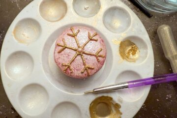 a pink macaron with a pink buttercream snowflake piped on top in the center of a paint palette and wells filled with gold luster dust suspension and a paintbrush