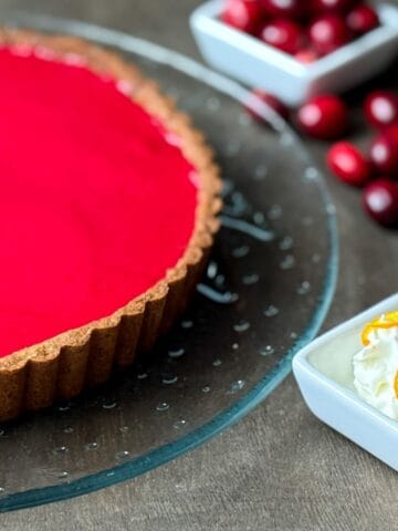 a Nut Free Cranberry Curd Tart on a clear glass platter with fresh cranberries around and a dish of whipped cream with a sugared cranberry and orange zest on top