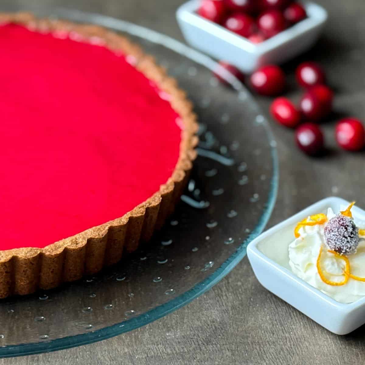a Nut Free Cranberry Curd Tart on a clear glass platter with fresh cranberries around and a dish of whipped cream with a sugared cranberry and orange zest on top