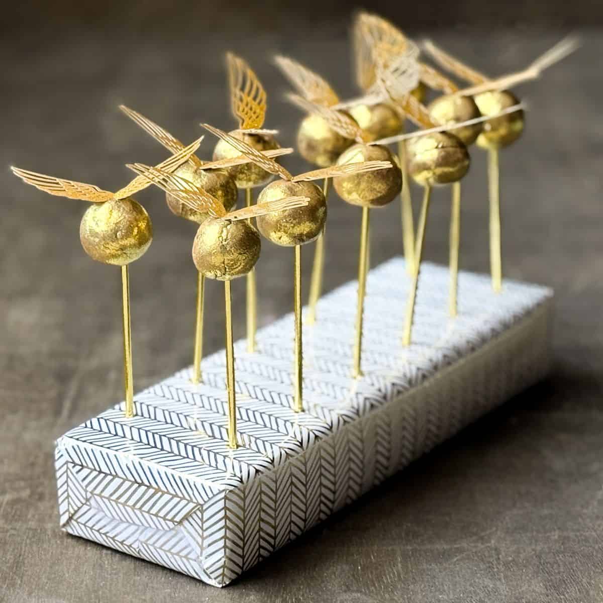 Golden Snitch Brigadeiros on gold sticks in a cake pop display block