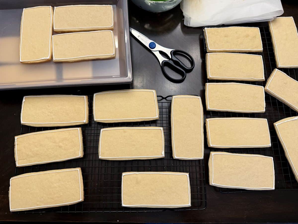 many cookies on cooling racks that have been outlined with white royal icing