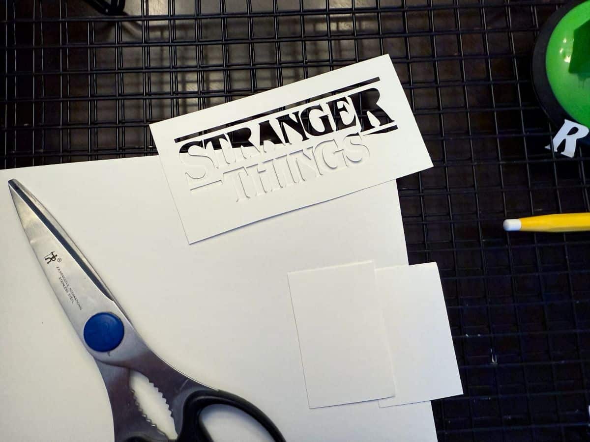 a white cardstock Stranger Things logo stencil with a pair of scissor and other pieces of cardstock