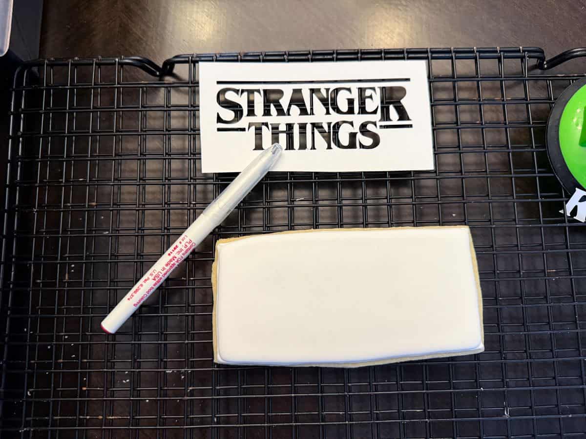 a white royal icing cookie next to a cardstock stencil of the Stranger Things logo and a red edible marker