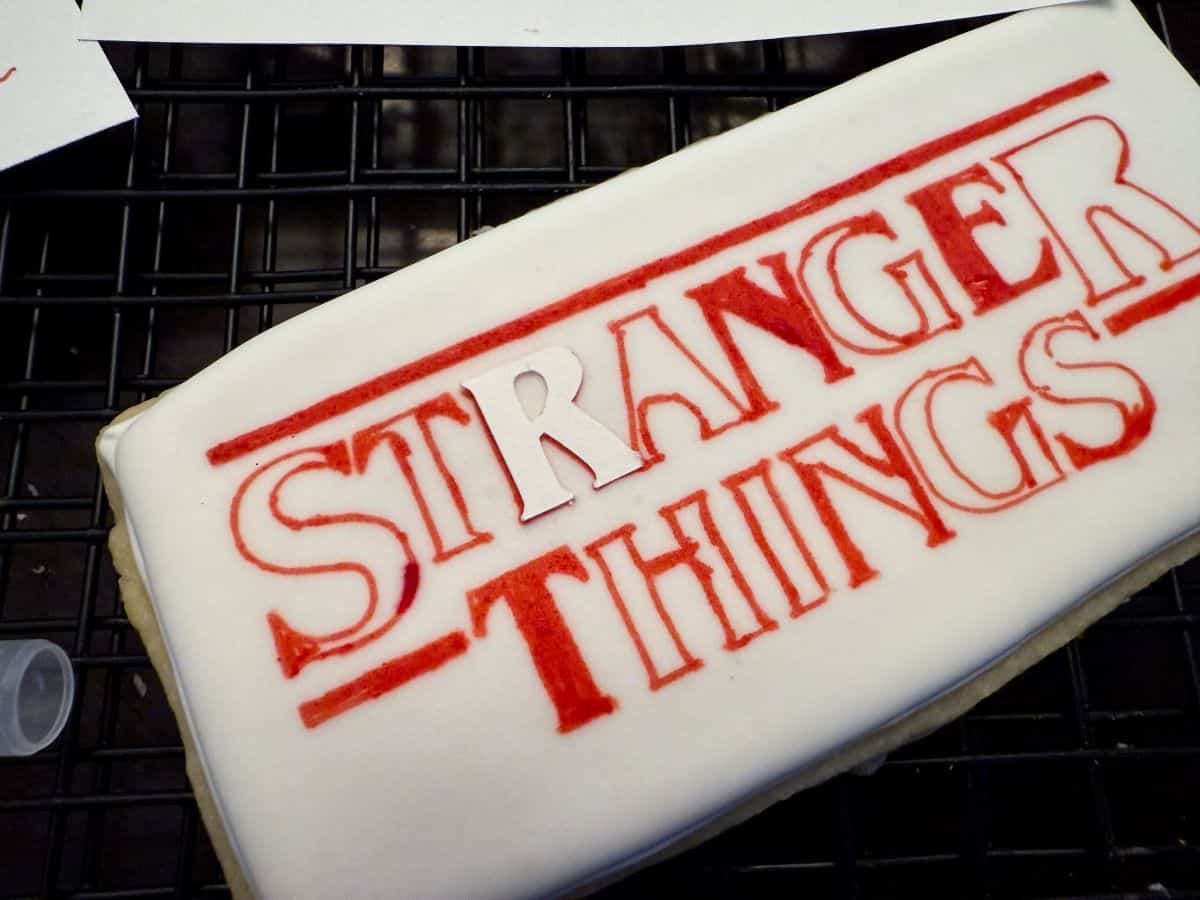 white royal icing cookie with red edible marker of the Stranger Things logo and a cardstock R to show how to draw the inside of the letter