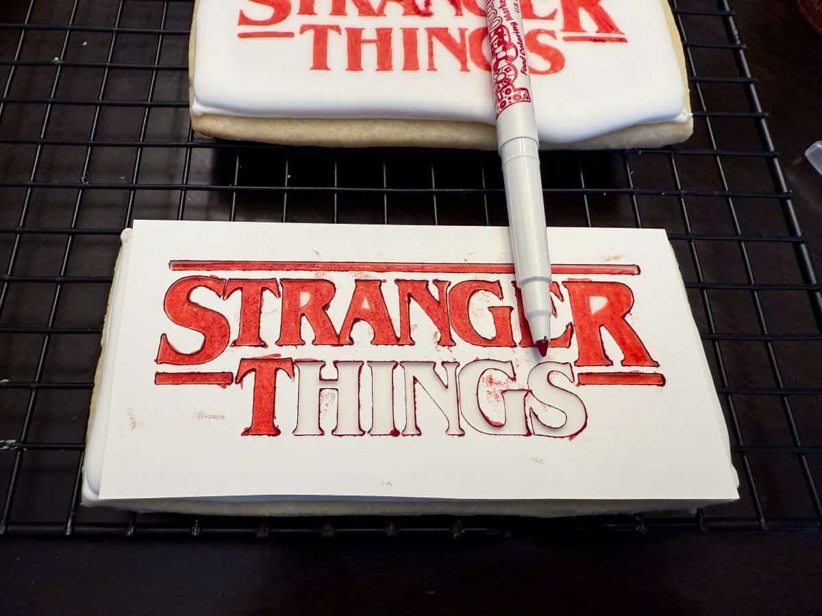 a cardstock Stranger Things stencil on top of a white royal icing cookie with half of the logo filled in with red edible marker