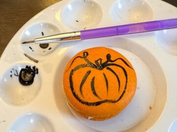 an orange macaron in the center of a paint palette with a pumpkin design painted on top with black food coloring gel
