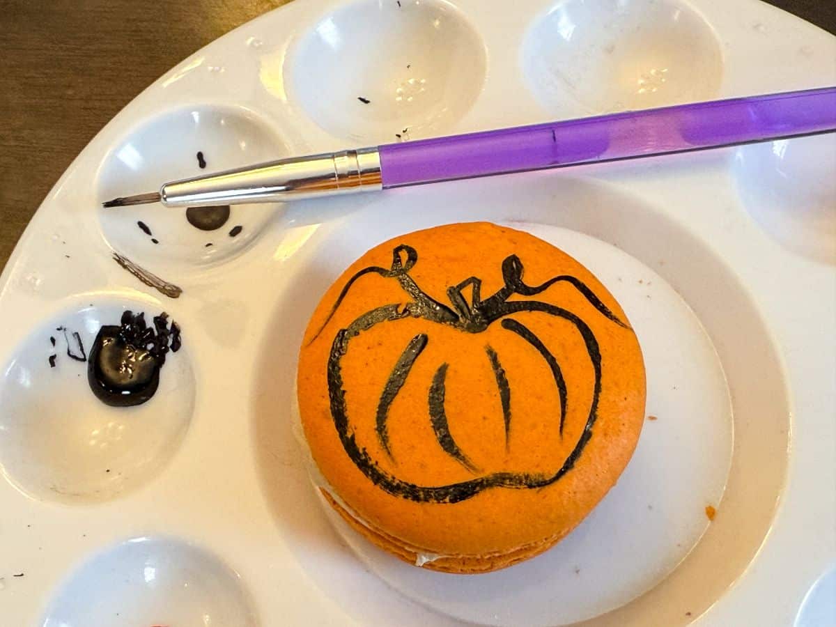 an orange macaron in the center of a paint palette with a pumpkin design painted on top with black food coloring gel